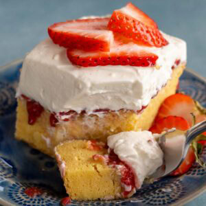 A big piece of strawberry poke cake with fluffy whipped topping and sliced strawberries on a blue plate