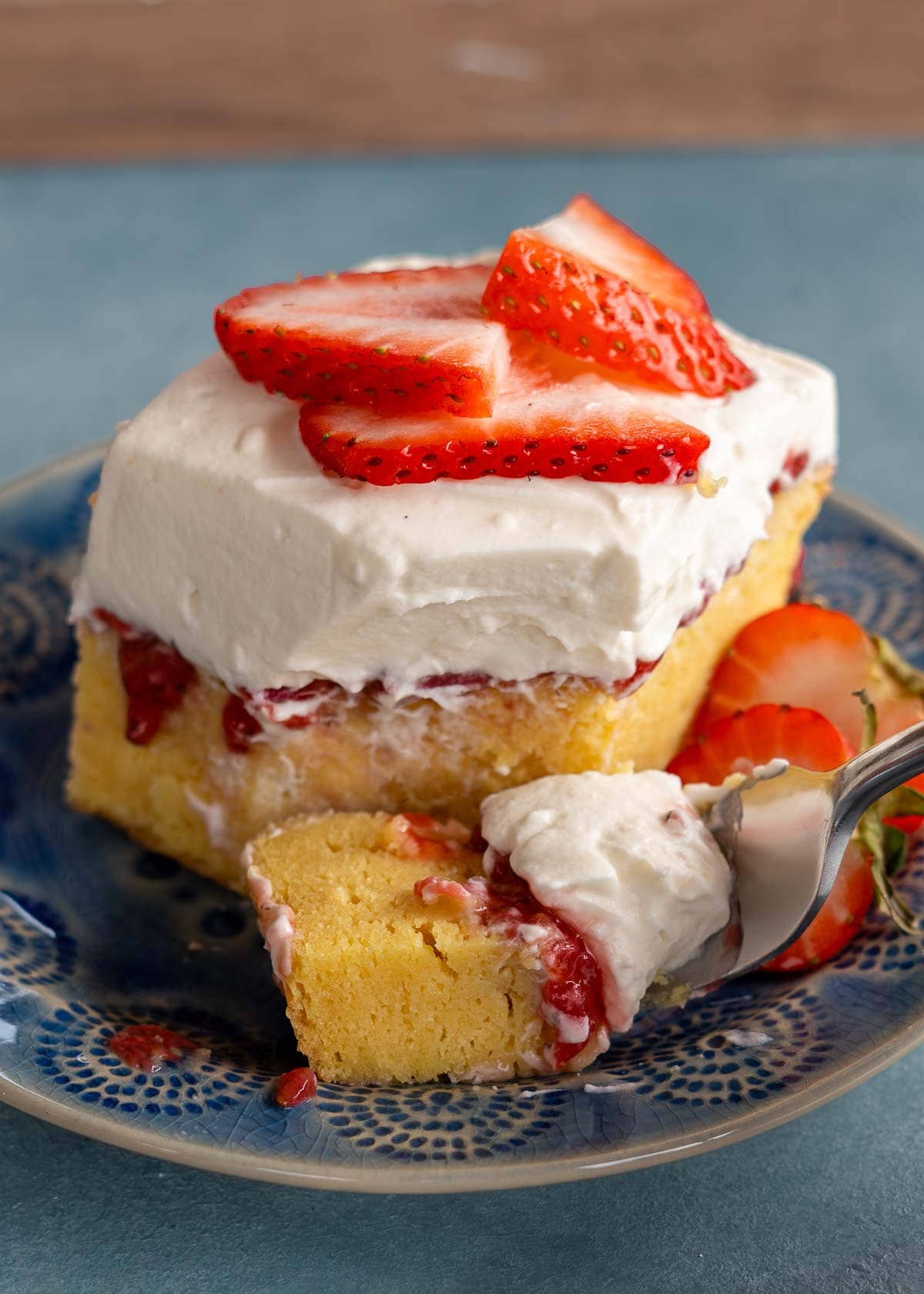 A big piece of strawberry poke cake with fluffy whipped topping and sliced strawberries on a blue plate